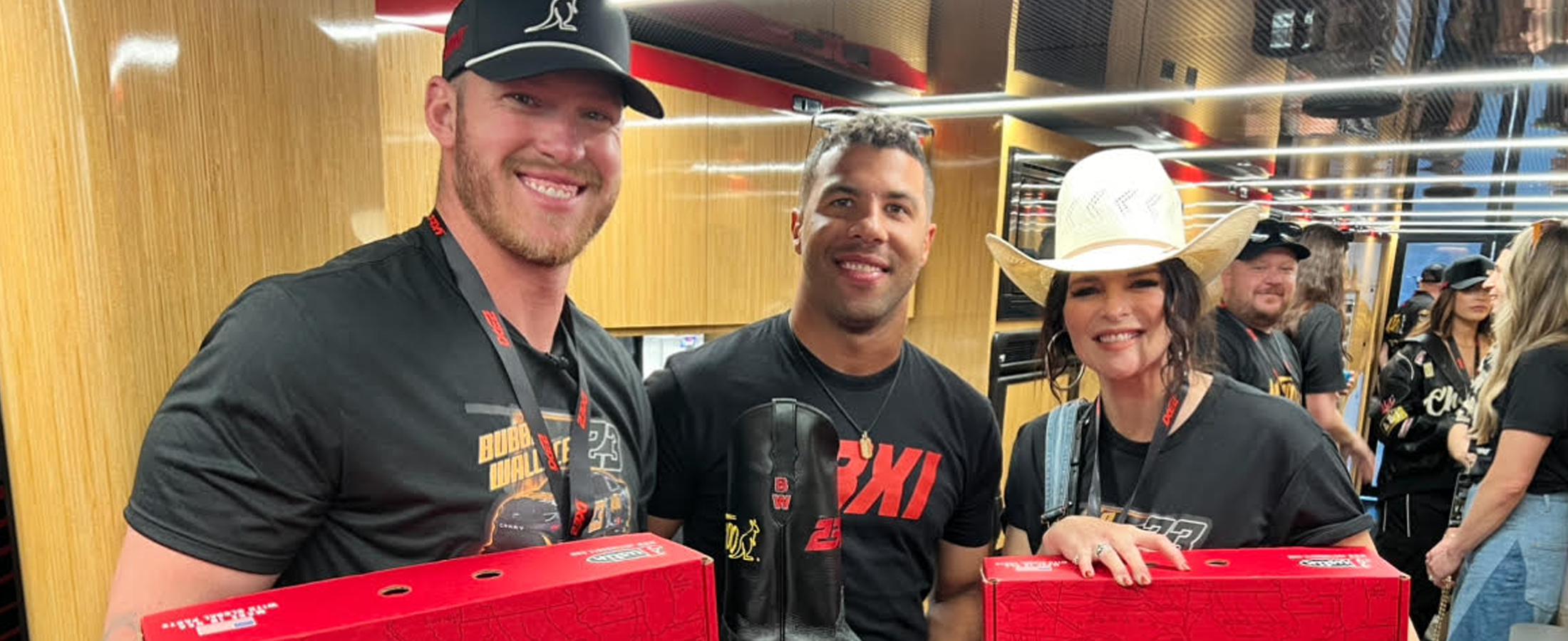 Alltroo Co-Founder Kyle Rudolph on the left holding a red Justin boot box, posing with 23XI Racing Driver Bubba Wallace, who is holding a black custom boot wearing a black t-shirt, standing next to Jenna Paulette on the right who is also holding a red Justin boot box. 